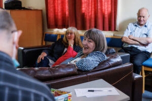 Ladies and gentlemen sit on sofas smiling while chatting in a Guideposts mental health support group.