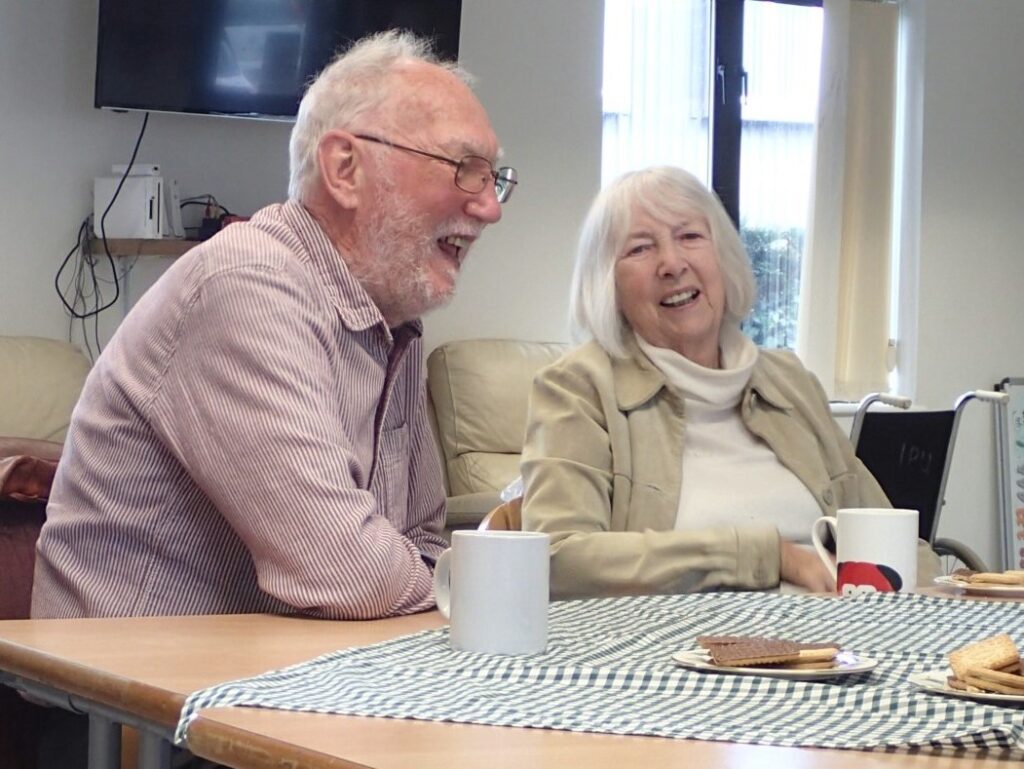 A laughing older couple sitting at a table with tea and biscuits.