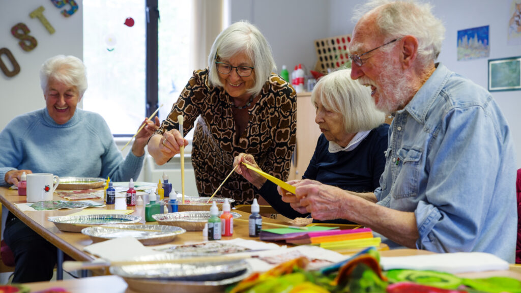 Four older Guideposts members sit at a table with paint, coloured paper, and brushes, smiling as they paint and chat.