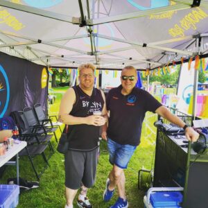 two men in shorts and t-shirts smiling to camera, standing next to a mixing desk, in a tent as in an outside broadcast. 