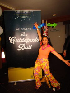 A smiling Guideposts staff member poses beside a sign reading 'The Guideposts Ball'.