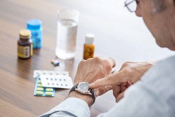 An elderly gentleman looks at his watch while sitting at a table. There are bottles of various tablets on the table.