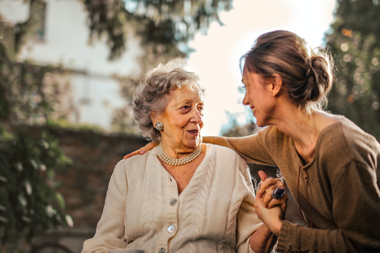 An older and a younger woman in a garden, looking at each other. The older woman has an expression of surprise and happiness, and the younger is smiling at her.