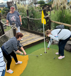 people playing a game of crazy golf - one woman pointing at the hole and another inexpertly pushing the ball towards it.  