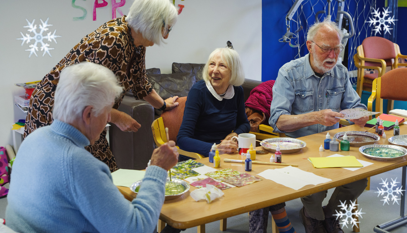 A group crafting around a table with paints. A woman in the middle is looking up gratefully, smiling, to look at the group facilitator. 