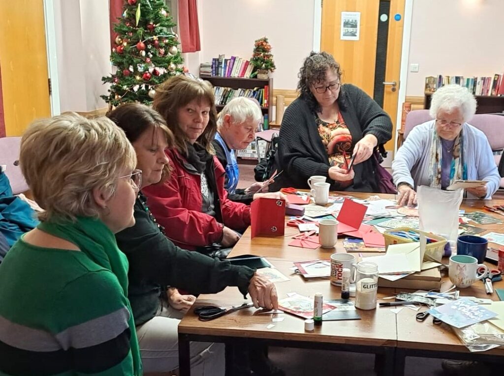 A group of 6 people making Christmas cards together.Most are concentrating on the materials, one is looking to the camera. Another is looking across the table.