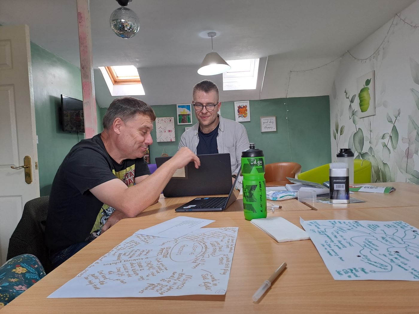 Two people working together at computers. with large pieces of paper on the table showing creative ideas