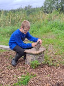 A boy in a blue jumper sitting on a bench in field, concentrating on moulding some clay