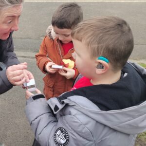 A boy wearing a grey jacket and a blue hearing aid is looking at the forest school leader who is handing him something small in a matchbox. There is another child in the background, looking excitedly into his matchbox.