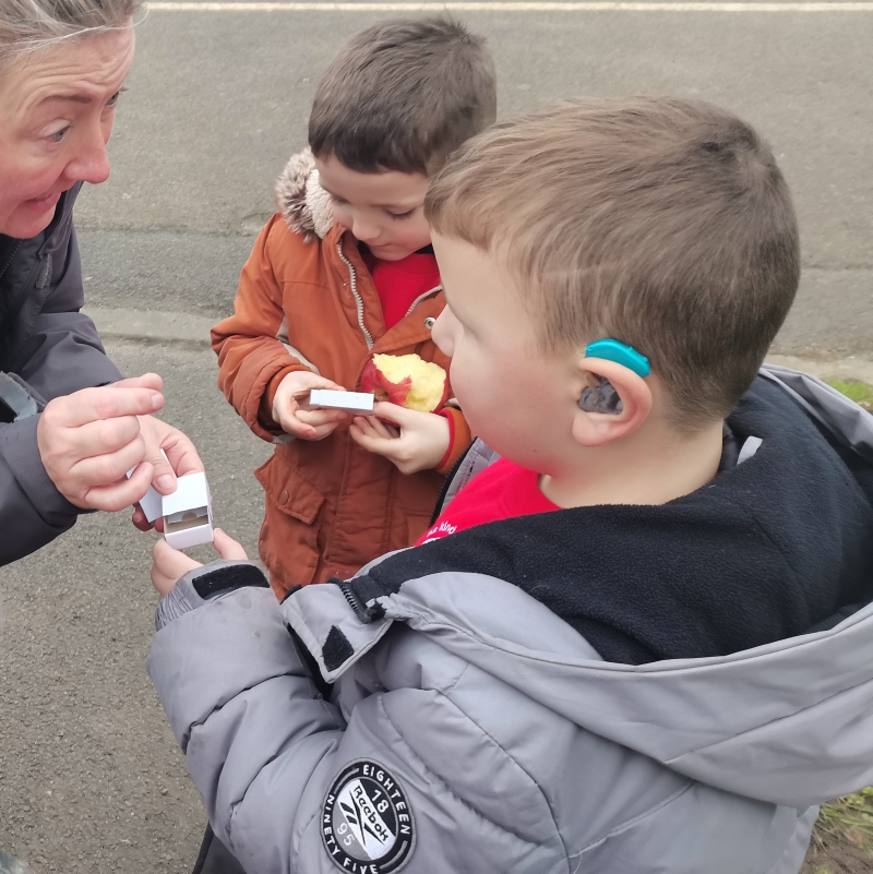 A boy wearing a grey jacket and a blue hearing aid is looking at the forest school leader who is handing him something small in a matchbox. There is another child in the background, looking excitedly into his matchbox. 