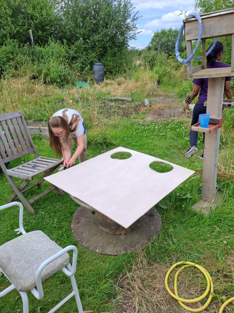 Person drawing on a large piece of pywood with 2 holes cut out of it, in a wild garden