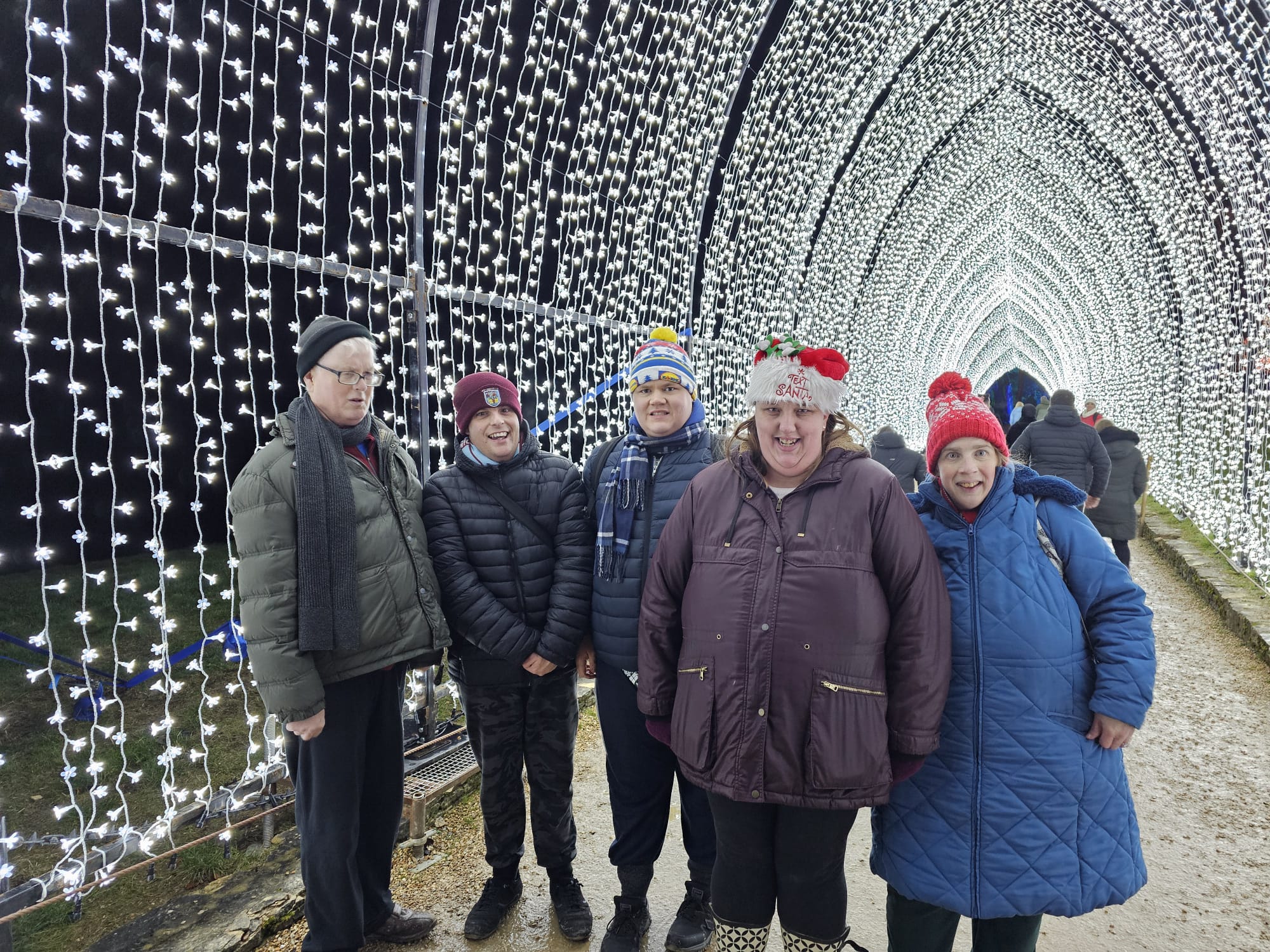 a group of 5 people standing inside a tunnel of fairy lights, looking at the camera in stunned amazement. Several are wearing red bobble-hats as well as coats and scarves