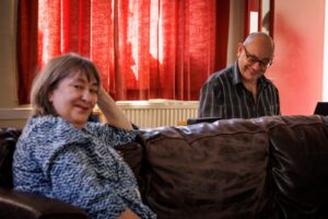 Woman leaning on back of sofa, looking at camera with a feint smile. Man sitting in background smiling