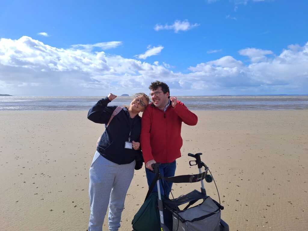 Two people on the beach, smiling with thumbs up. One of them has a walking frame. It is a beautiful day with blue sky and a vast beach