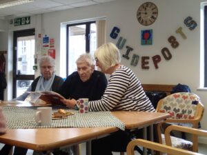 Three people at a table with tea and biscuits. The closest lady is talking the older man next to her, looking at some pictures.