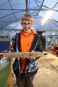 boy in orange jumper, smiling excitedly and holding a giant wooden pencil