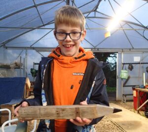 boy in orange jumper, smiling excitedly and holding a giant wooden pencil