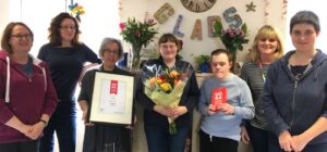 Group of people posing to camera. In the middle are three people, holding flowers, a certificate and a trophy.