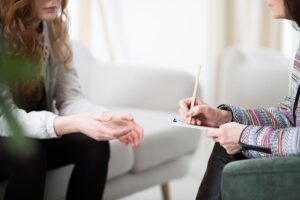 Two women sitting on sofas opposite each other. The faces are not visible but the hands show signs of serious discussion. One has a clipboard and pencil.