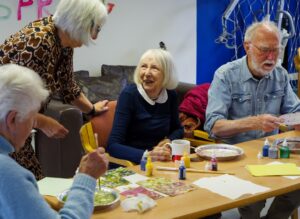 A lady sat at a table wearing in a blue sweater and white collar with white hair is looking up and smiling at the lady standing next to her, with bright eyes.