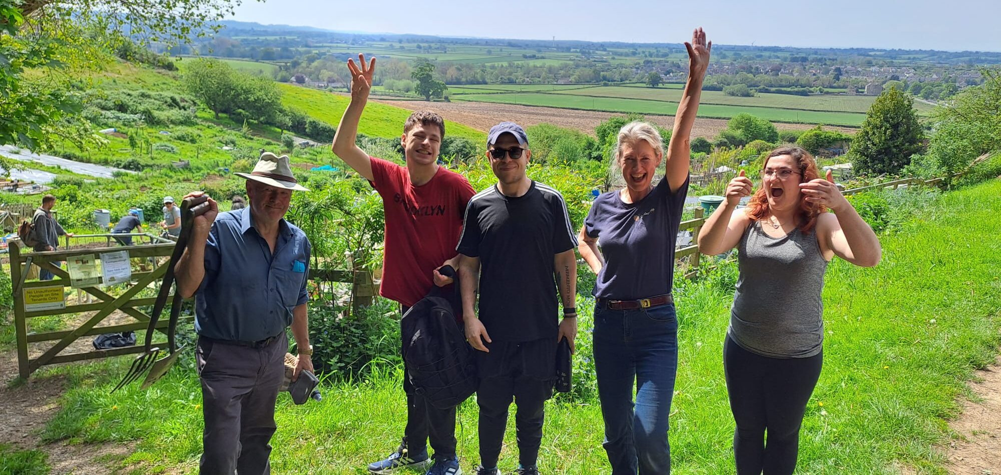 5 people standing in front of an amazing view over green english countryside. Their expressions are celebratory and 2 of them have hands up in the air. One is carrying a garden fork, and just behind them is an allotment site with people working in it.