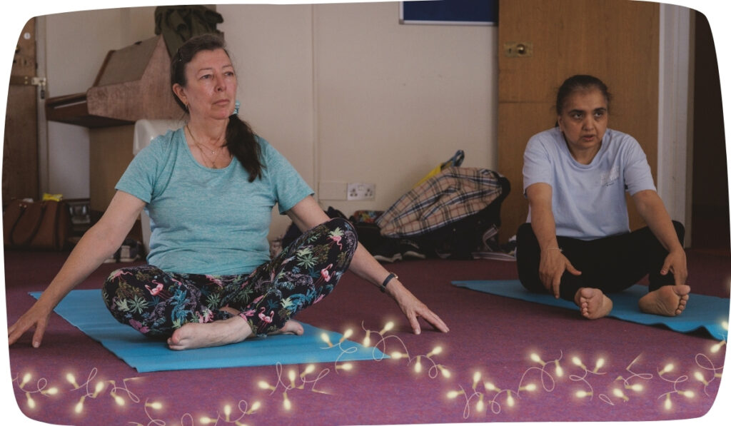 two women in a yoga class, with fairy lights around them. One woman is sitting cross-legged and looking into the distance. The other is sitting and reaching towards her toes.