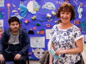 a woman is standing and a younger man sitting, in front of a colourful wall of sea art. The woman is looking to camera and smiling. The man is looking at her, with a neutral expression.