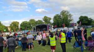 people watching a band on a stage at a music festival. There is a wide range of people, and 2 in the front wearing hi-viz vests.