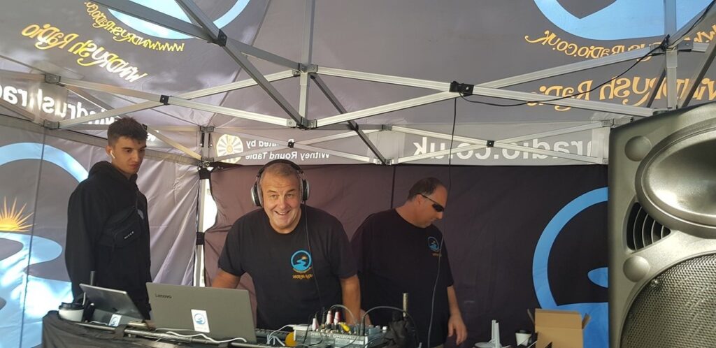 photo of 3 men behind a mixing desk, in an outdoor broadcast tent. The man in the middle is looking to the camera and grinning.