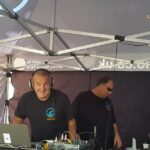 photo of 3 men behind a mixing desk, in an outdoor broadcast tent. The man in the middle is looking to the camera and grinning.