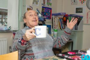 A man posing for the camera, smiling and with hands up, as if to say "cheers" with a coffee cup.
