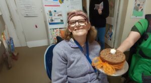 A woman sitting, with a cake held in front of her. The cake has chocolate icing and a single candle on it.