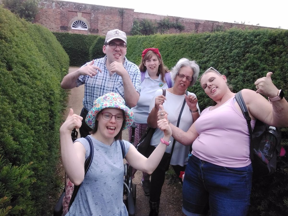 a group of people in a maze, with hedges all around them. They all have thumbs up and looking very happy. They are wearing summer clothes, one wearing a cap and anther a cloth hat.