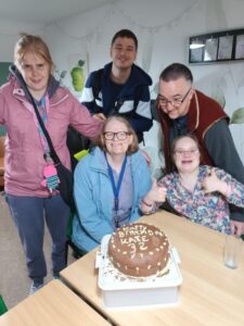 A group of 5 people around a birthday cake. The cake has "Happy Birthday Kate 32" written in white chocolate on a dark chocolate icing. Everyone is looking happy and celebratory.