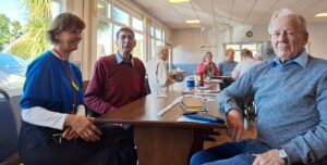 Three people sitting at the end of a long table, with a game of dominos. The man on the right is looking to camera with a content but unsure gaze. A woman wearing an identity badge is looking at him encouragingly, and the second man is looking cheerfully between them.