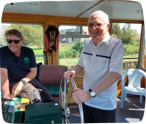 an older man holding the steering wheel of a river boat and smiling. The boat driver is sitting nearby.