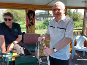 an older man holding the steering wheel of a river boat and smiling. The boat driver is sitting nearby.
