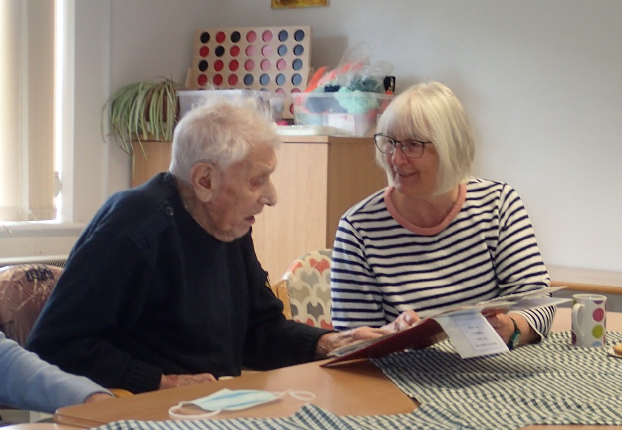 An elderly man and woman are looking at each other in discussion, holding some paperwork on a clip-board. There is a checked table cloth and cup of tea on the table.