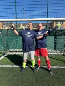 two young men in front of a football goal, wearing Guideposts Trust shirts and goalie gloves. They are smiling and have thumbs up to the side. 