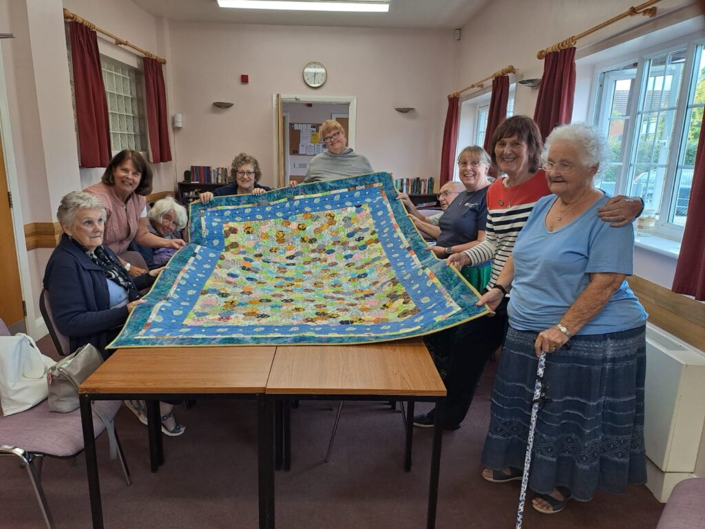 group of people around a table, holding a large colourful patchwork quilt between them