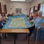 group of people around a table, holding a large colourful patchwork quilt between them
